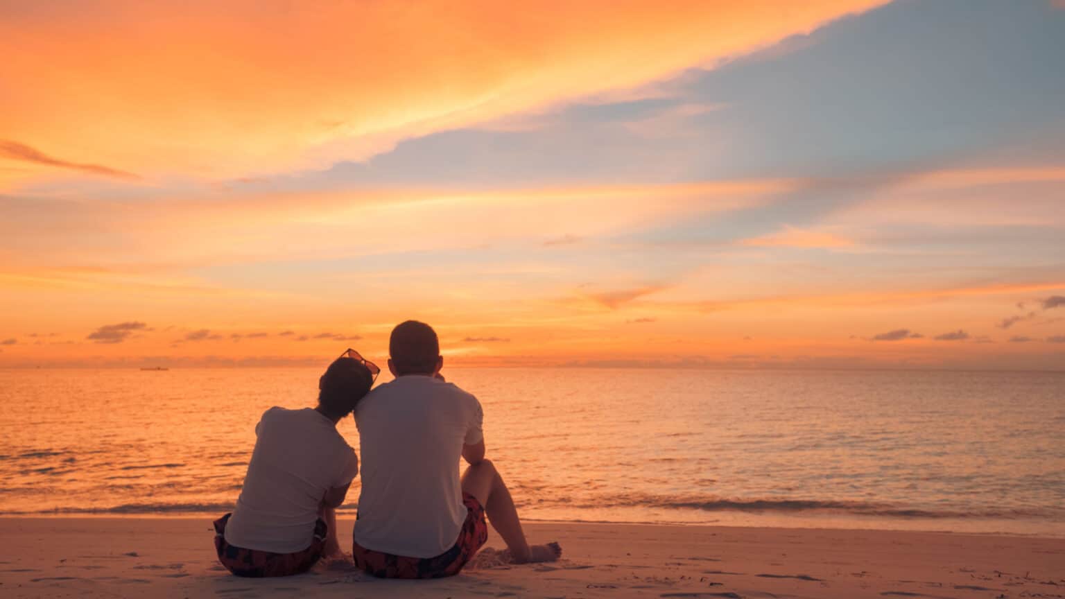 Couple in love watching sunset together on beach travel summer holidays. People silhouette from behind sitting enjoying view sunset sea on tropical destination vacation. Romantic couple on the beach