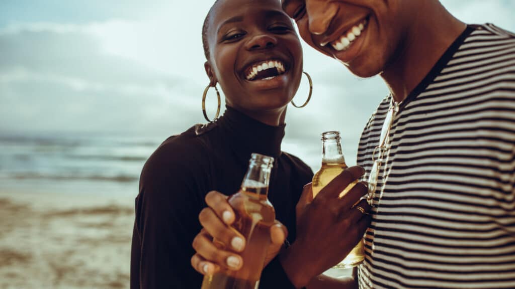 African couple on vacation having beers at the beach. Cheerful young man and woman on summer holidays having beers and smiling at the beach.